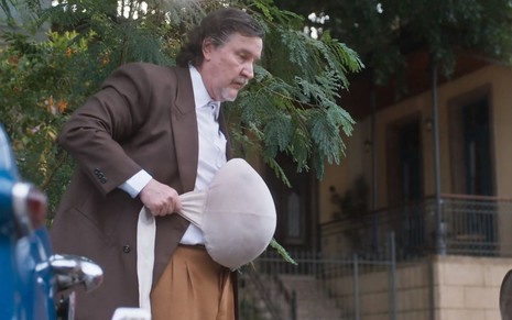 O ator Antonio Calloni como Matias em Além da Ilusão; ele está olhando para baixo com cara séria enquanto segura em frente a sua barriga uma falsa barriga de grávida