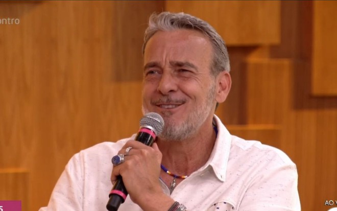 O ator Alexandre Borges dá um sorriso envergonhado enquanto segura o microfone. Ele veste uma camisa branca, pulseiras e dois anéis na mão direita. No fundo, o cenário do Encontro.