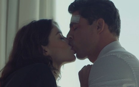 Alinne Moraes e Cauã Reymond dando um beijo em uma cena de sexo da novela Um Lugar ao Sol. Ela está de camisa preta, ele de camisa branca quadriculada e com um curativo na cabeça.