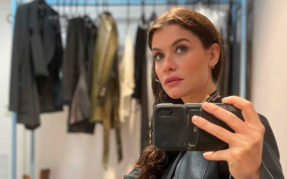 A atriz Alinne Moraes tira uma selfie diante de um espelho com uma arara de roupas no fundo nos Estúdios Globo, no Rio