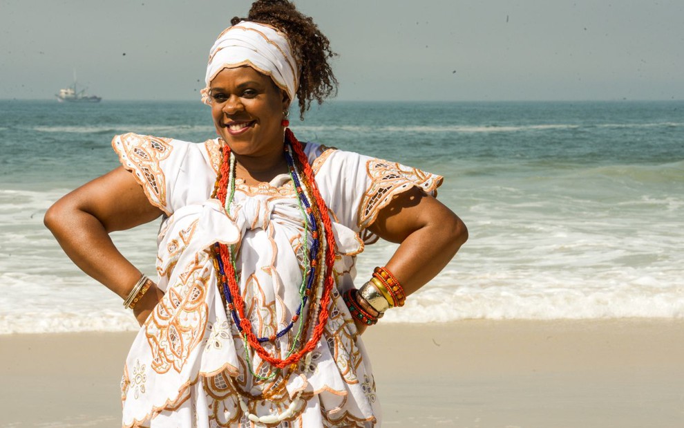 Cacau Protásio está caracterizada com roupas de umbanda em uma praia e sorri para a câmera com os dois braços na cintura