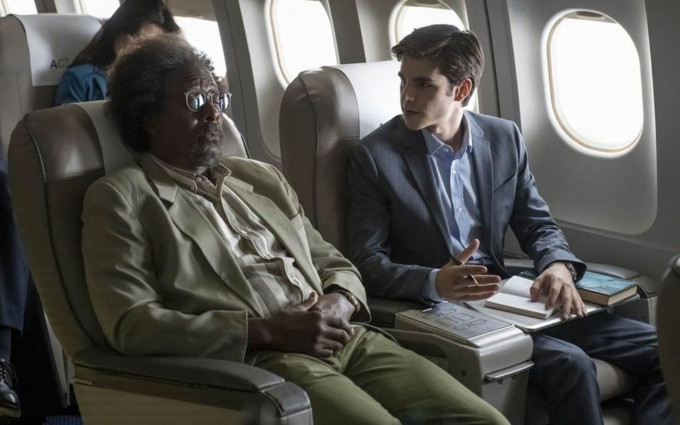 Clarke Peters e Álvaro Mel estão sentados em poltronas de um avião. O primeiro está tentando descansar, enquanto o segundo faz perguntas