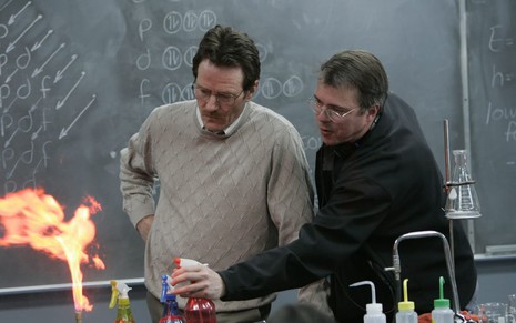 Bryan Cranston e Vince Gilligan estão no cenário de uma sala de aula, com uma chama acesa na frente deles