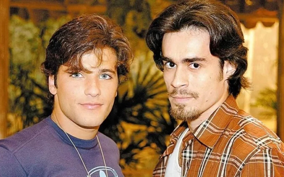 Os atores Bruno Gagliasso e Erom Cordeiro lado a lado, olham para a câmera com expressões sérias, em cenário da novela América