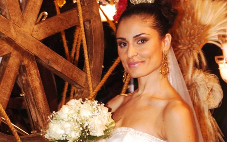 A atriz Franciely Freduzeski vestida de noiva, com coque e véu no cabelo, segura buquê de flores em cena da novela América
