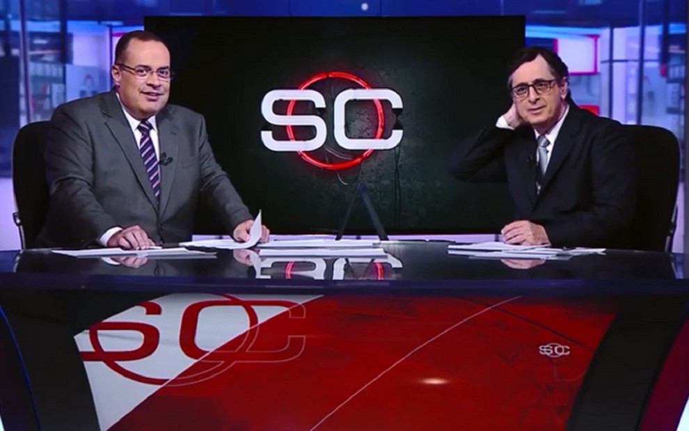 Paulo Soares, o Amigão, e Antero Greco, nos estúdios da ESPN Brasil. Amigão usa um terno cinza e Antero Greco veste uma peça preta.