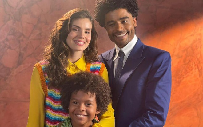 Com blusa amarela e colete de tricô listrado e colorido, Camila Queiroz posa ao lado de Diogo Almeida, que está de terno azul, e abraçada a Levi Assaf
