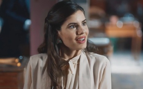 A atriz Camila Queiroz com expressão de curiosidade, camisa bege e cabelo preso em cena de Amor Perfeito
