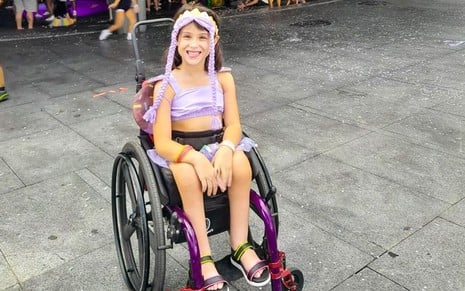 Vitoria Pabst está em sua cadeira de rodas, sorrindo, usando uma tiara com tranças lilás