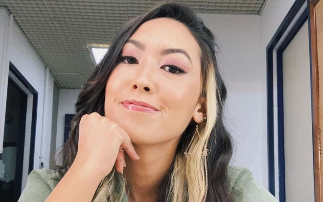 Com mechas loiras e blusa verde, atriz asiática Ana Hikari sorri de boca fechada com o rosto apoiado no cotovelo