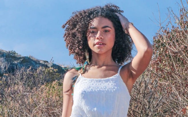 A atriz Alana Cabral mexe no cabelo e usa vestido branco de alcinha ao posar para fotos em uma praia