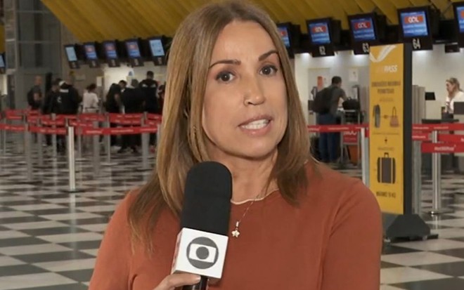 Ananda Apple no SP1 desta sexta-feira (13), ela está em um aeroporto com um microfone da Globo na mão e veste uma blusa vermelha