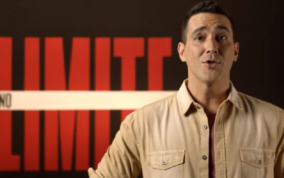 André Marques com uma camisa marrom falando sobre o No Limite, com o logotipo do programa ao fundo em um painel preto