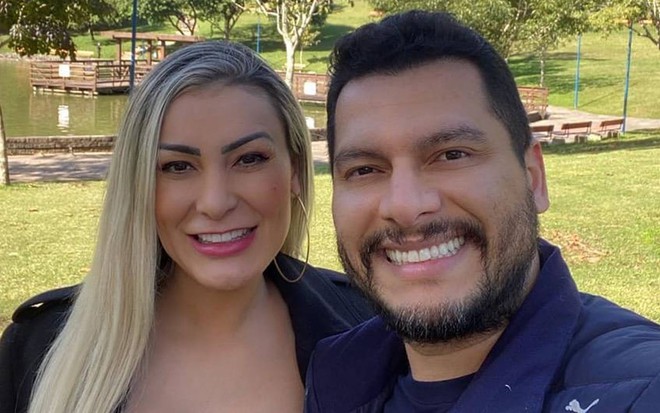 Andressa sorri, veste blusa preta e está ao lado de Thiago; ele sorri e veste jaqueta azul