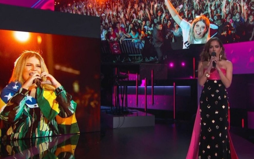 No palco do Grammy Latino, Anitta segura um microfone; imagens de Marília Mendonça aparecem nos telões