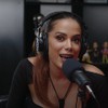 Anitta com um fone de ouvido na cabeça e um microfone a sua frente durante entrevista ao Poddelas