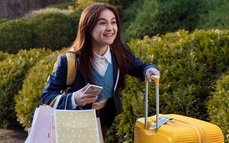 Anna Cathcart usa uniforme escolar e empurra uma mala de viagem amarela