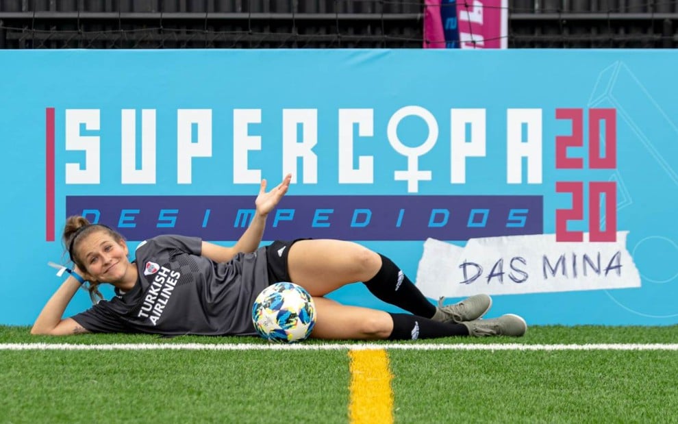 Apresentadora Alê Xavier deitada em campo com meião e calção preto e camisa cinza com uma bola na frente e placa colorida atrás na Supercopa das Mina 2020
