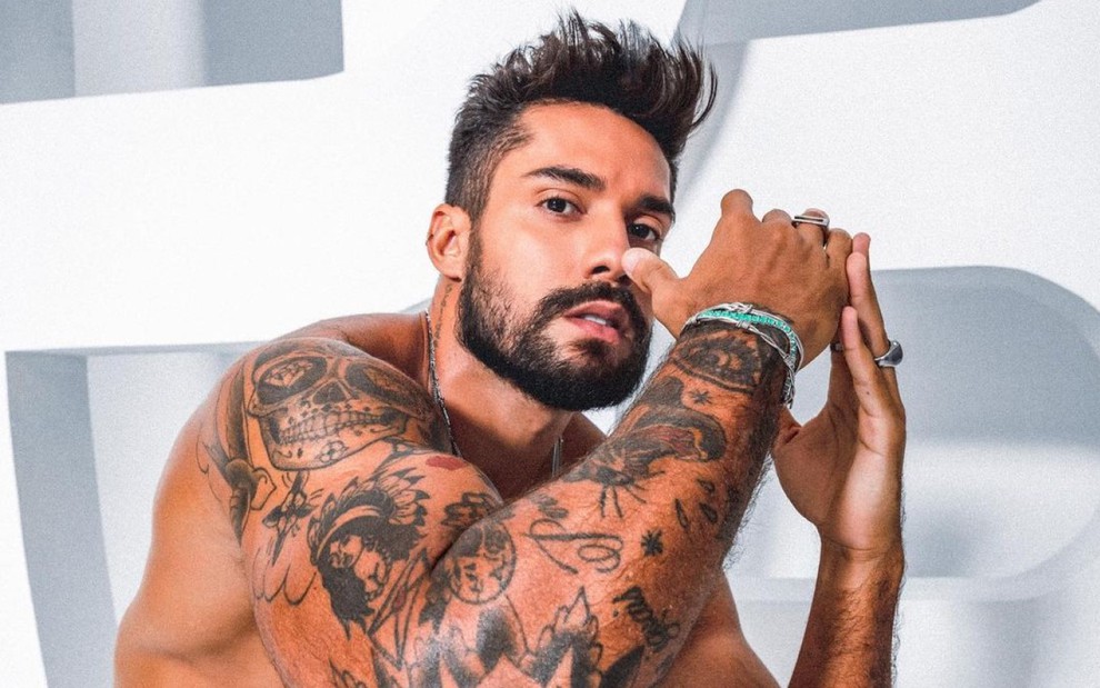Arcrebiano de Araújo exibe suas tatuagens sem camisa, olhando de lado e com os dois braços levantados em direção ao rosto