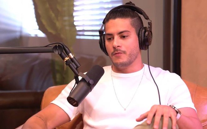 Arthur olha para baixo, veste camiseta branca e usa fones de ouvido preto