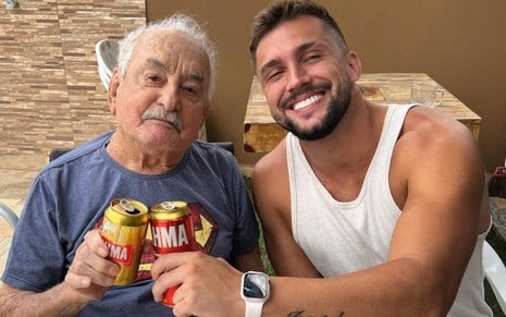 Arthur Picoli e seu avô estão sorridentes e brindam latas de cerveja