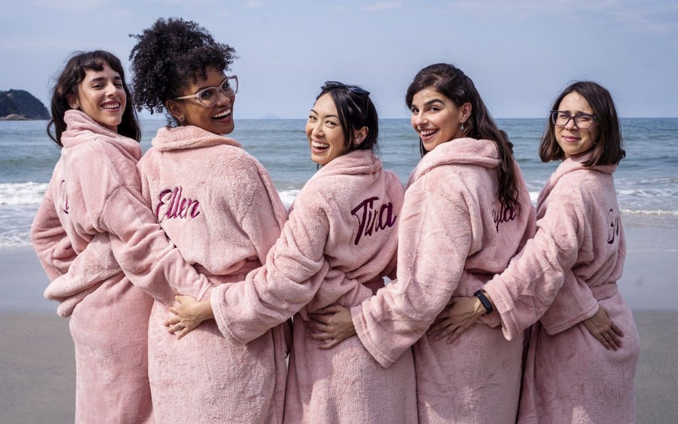 As cinco protagonistas de As Five lado a lado, de costas, abraçadas, sorriem para a câmera, usam roupões cor de rosa em uma praia