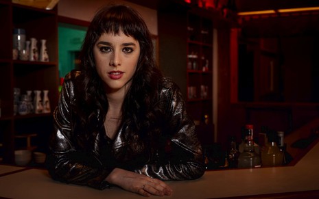 Vestida com jaqueta preta em cima de uma blusa da mesma cor, Manoela Aliperti posa para a foto no cenário de um bar