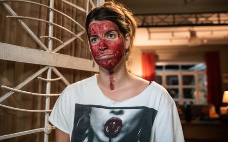 A atriz Maria Bopp com máscara vermelha no rosto, expressão séria, camiseta preta e branca, em cena da série As Seguidoras