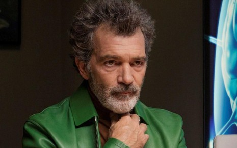 Salvador (Antonio Banderas) de blusa marrom e jaqueta verde com a mão direita no pescoço olhando para a direita em cena do filme Dor e Glória