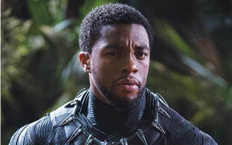 T'Challa (Chadwick Boseman) com armadura preta e detalhes em prata com olhar sério com mata ao fundo desfocada em cena do filme Pantera Negra