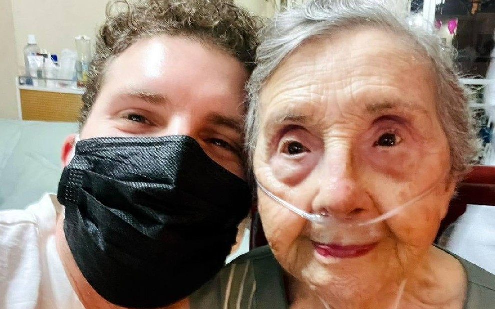 Thiago Fragoso no lado esquerdo de máscara preta e sua avó Edith no lado direito com tubos de respiração no nariz com rostos colados
