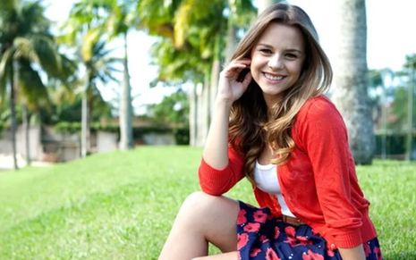 Daniela Carvalho em 2010. Ela posa sentada em gramado, de saia florida, casaco vermelho, cabelos soltos e sorrindo