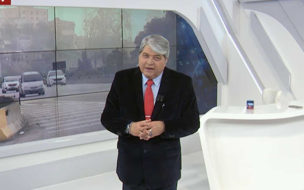 O apresentador Datena com um terno preto, camisa azul e gravata vermelha no centro do cenário do Brasil Urgente