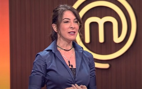 Ana Paula Padrão está de roupa azul no estúdio do MasterChef; o logo do reality culinário aparece ao fundo da imagem