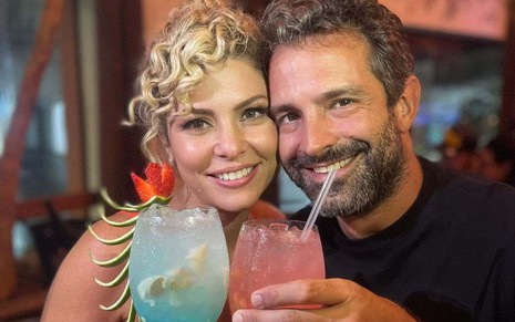 Bárbara Borges e Iran Malfitano com os rostos coladinhos, sorrindo e segurando drinks