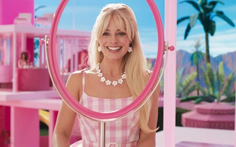Margot Robbie em cena como a protagonista de Barbie, de vestido rosa e branco, em frente a aro rosa, no cenário do mundo de Barbie