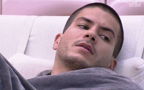 Arthur está sentado em um sofá branco e está coberto com um cobertor cinza