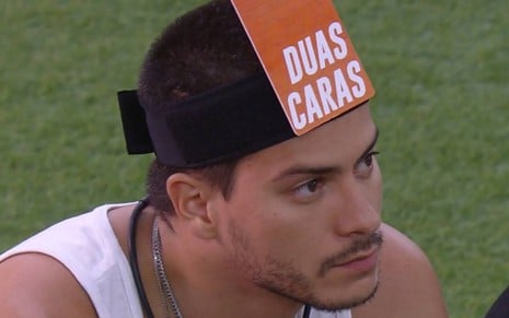 Arthur veste regata branca e está com uma faixa preta na cabeça com uma placa "duas caras" colada