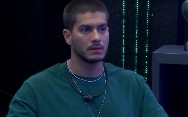 Arthur olha para TV e veste camiseta verde