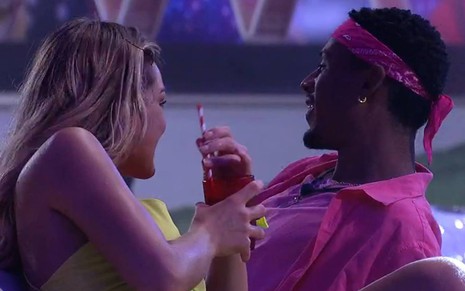 Bárbara está sentada, olha para Paulo André e veste um vestido amarelo; Paulo André usa camiseta rosa e lenço rosa na cabeça