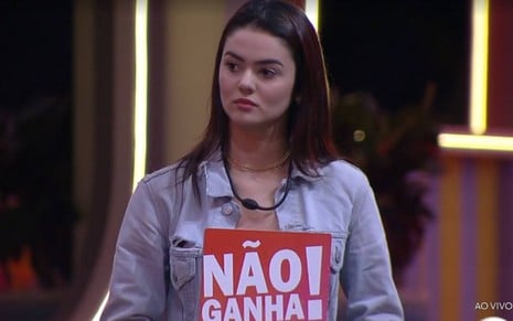 Eslô veste top branco e jaqueta jeans; ela segura a placa "não ganha"