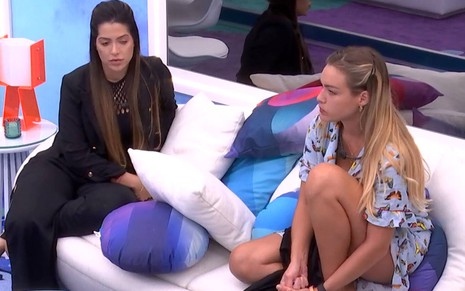 Laís veste conjunto preto e está sentada em um sofá branco ao lado de Bárbara; Bárbara veste pijama azul e está com o cabelo solto