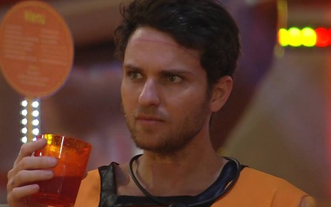 Lucas segura um copo, veste camiseta laranja e olha para o lado no reality show da Globo