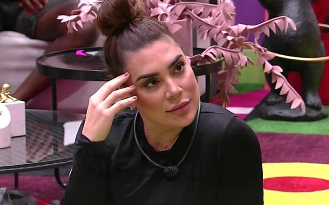 A participante Naiara Azevedo no BBB 22, de blusa preta, expressão séria, coque no cabelo e mão na testa