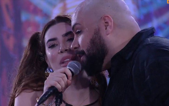 Naiara canta com Tiago; o apresentador segura um microfone e veste camiseta preta