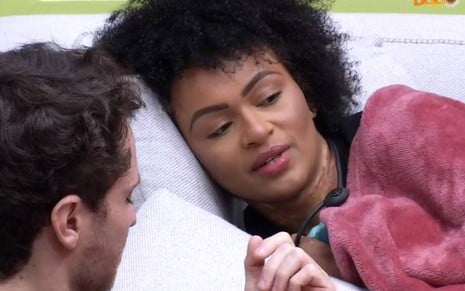 Lucas olha para Natália; Natália está deitada e coberta por um cobertor rosa