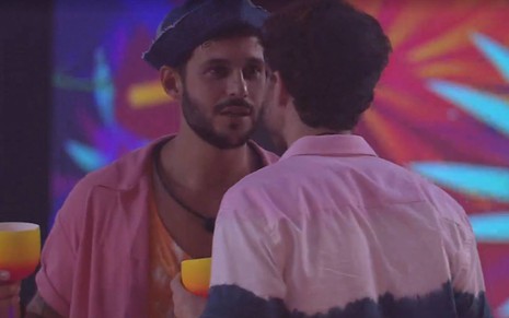 Rodrigo veste camiseta laranja com camisa rosa, usa chapéu na cabeça e olha para Lucas; Lucas veste camiseta rosa com detalhe preto e está de costas
