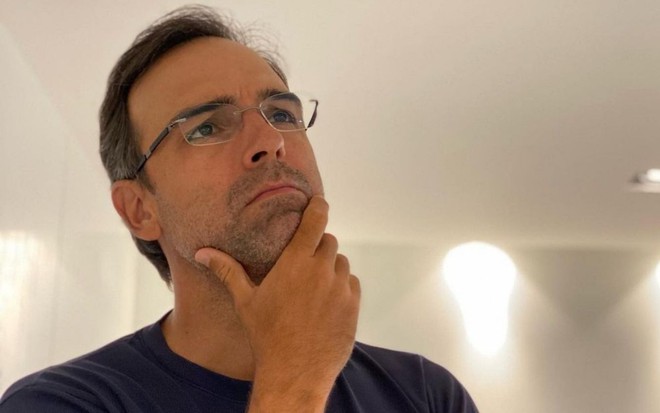 Imagem de Tadeu Schmidt de camiseta preta e óculos, com a mão no queixo