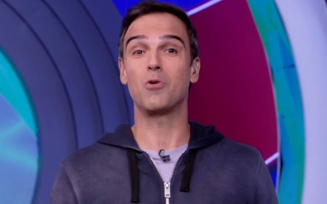 Tadeu está na frente de um fundo azul, veste jaqueta azul e camiseta cinza no estúdio do BBB 22
