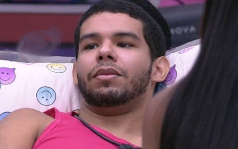 Vinicius está deitado, veste regata rosa e usa microfone no pescoço